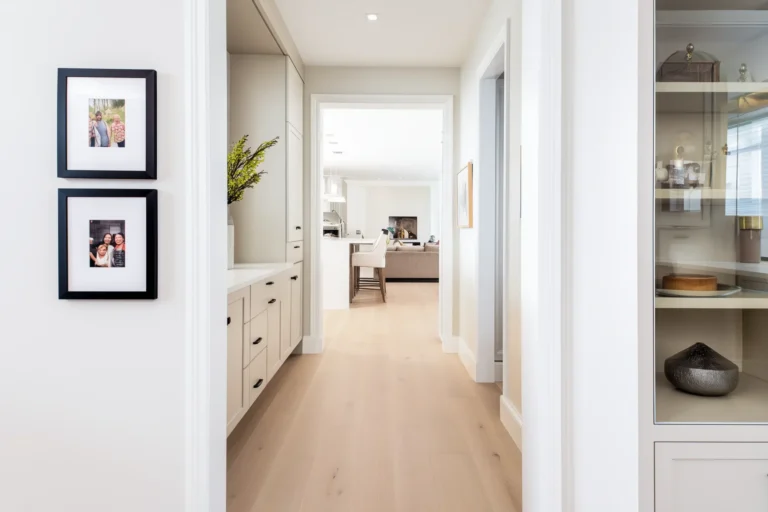 A bright, modern hallway with light wood floors leading to an open-concept kitchen and living area, featuring built-in cabinetry and framed photos on a white wall.