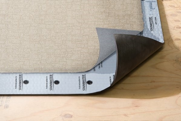 Layers of flooring installation on a plywood subfloor, showing a beige textured carpet peeled back to reveal a branded Stainmaster carpet cushion underlayment.